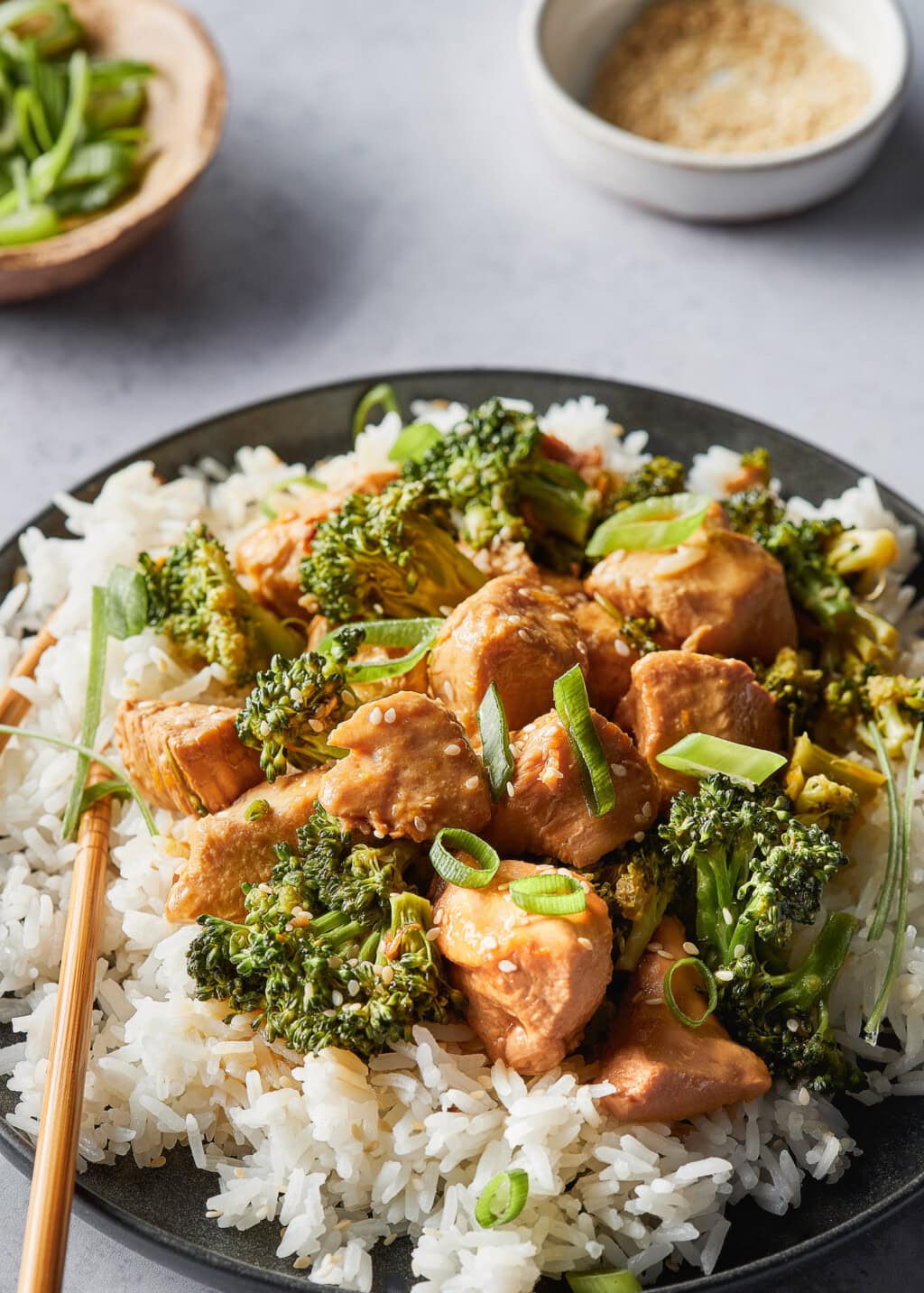 A close-up shot of a black plate filled with white rice, topped with tender, golden-brown chicken pieces coated in a glossy orange sauce, and bright green broccoli florets. The dish is garnished with sliced green onions and sesame seeds. Bamboo chop sticks rest on the edge of the plate. A small bowl, filled with chopped greens sits in the background.