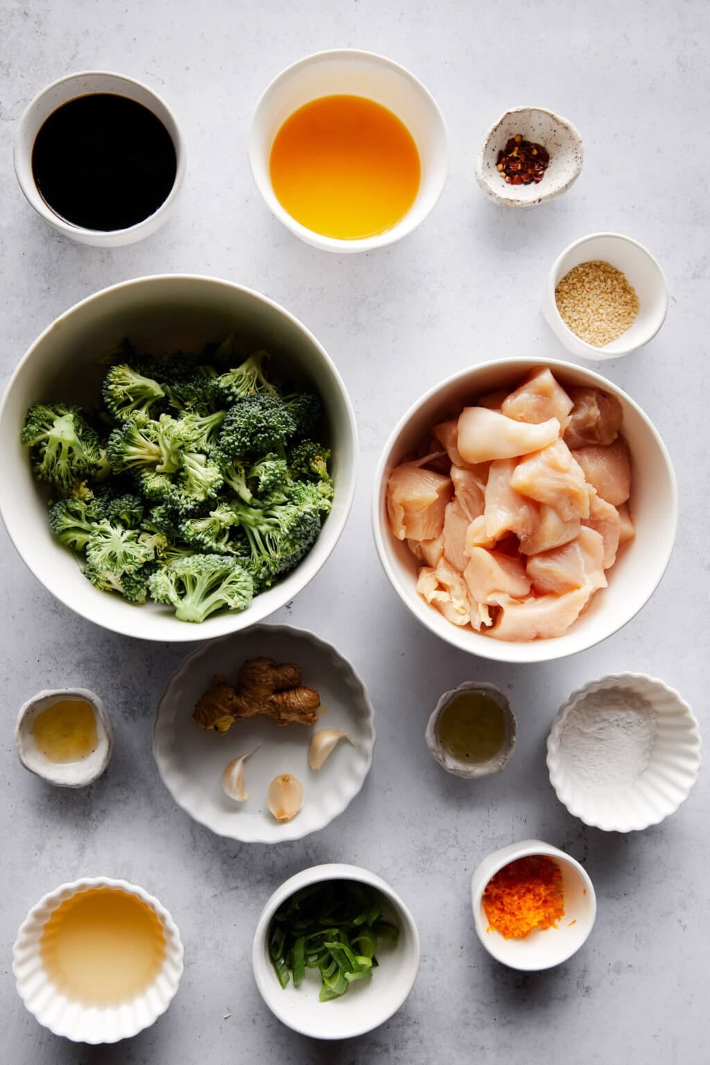Overhead view of various ingredients for Instant Pot Orange Chicken, neatly arranged in individual bowls on a light gray surface. Ingredients include chicken cubes, broccoli, orange juice, soy sauce, sesame seeds, ginger, garlic, green onions, rice vinegar, sesame oil, crushed red pepper, and arrowroot starch.