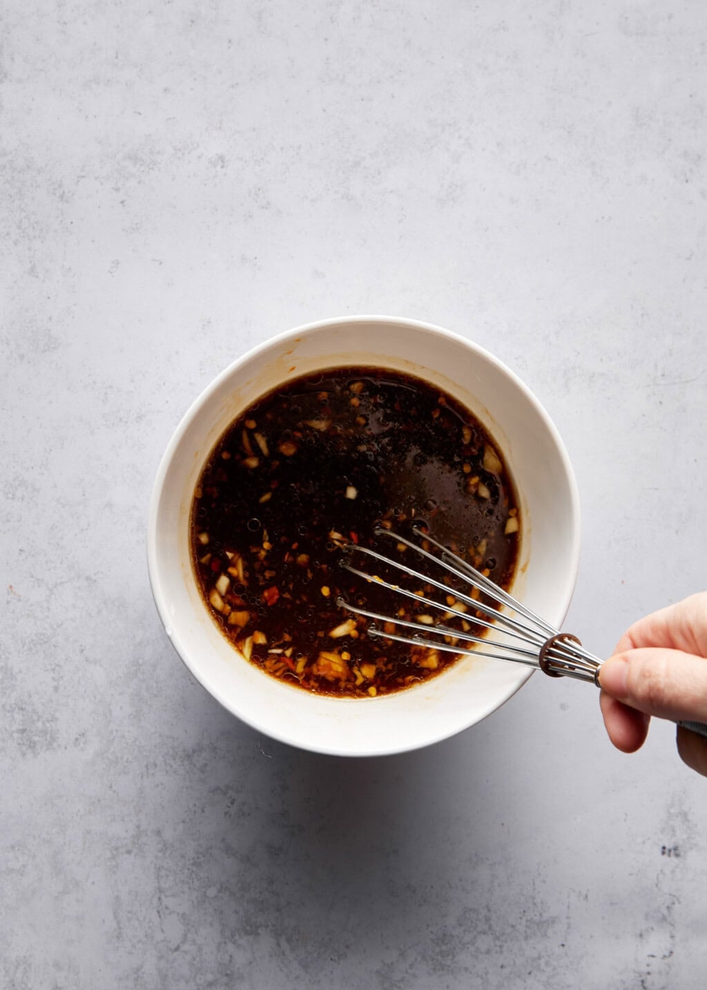 Overhead view of a hand whisking a dark brown sauce in a white bowl. The sauce contains small pieces of garlic, ginger, and other spices. The bowl is centered on a light gray surface. The hand holding the whisk is visible in the lower right corner.