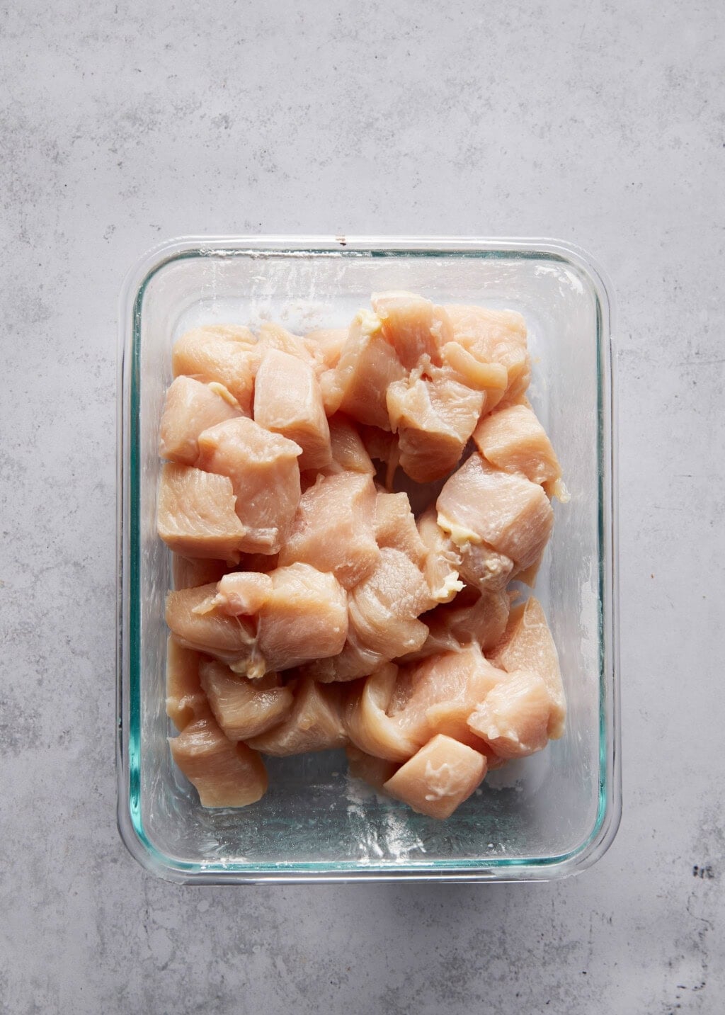 Overhead view of cubed chicken coated in arrowroot starch inside a clear glass container. The chicken pieces are light pink and evenly coated with a white powder, the arrowroot starch. The container has a light blue rim and is placed on a gray surface.