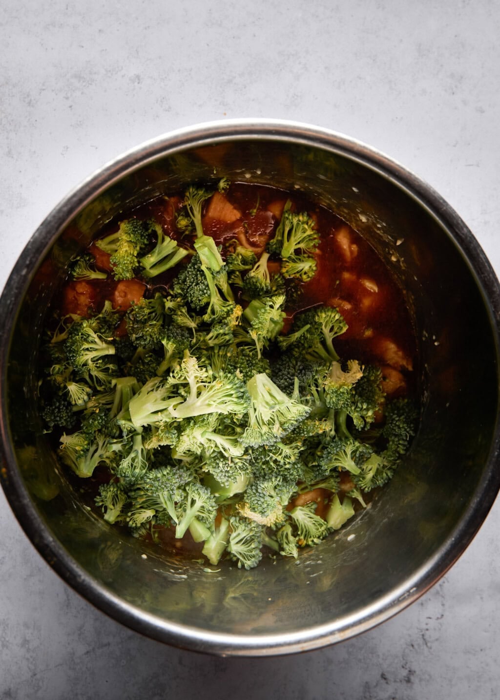 A top-down shot shows an Instant Pot filled with orange chicken and broccoli. The bottom of the pot contains orange chicken in a dark sauce and is coated with broccoli.