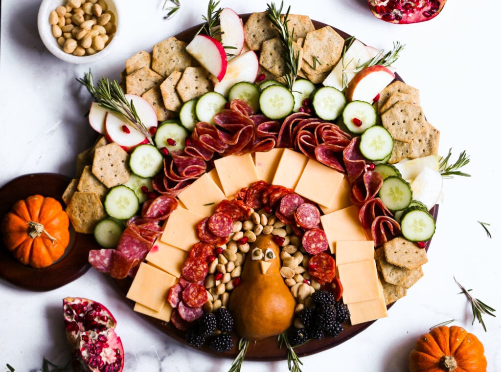 Festive turkey charcuterie board with cheddar, gouda, salami, crackers, cucumber, fruit, and nuts. A fun Thanksgiving appetizer idea.