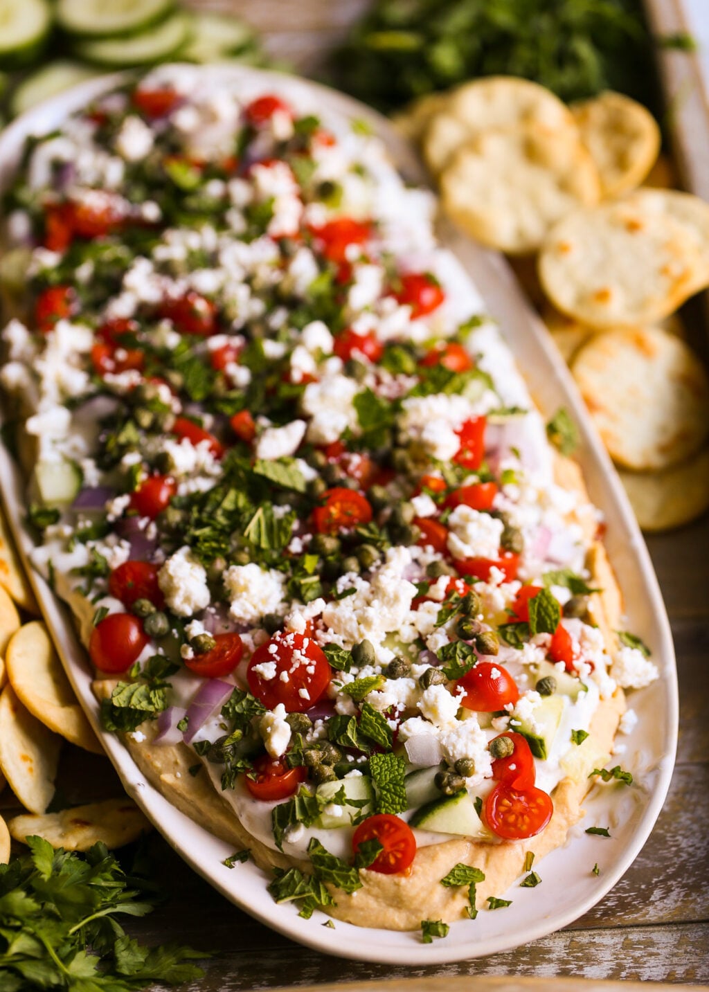 Easy layered Greek dip recipe with hummus, tzatziki, cherry tomatoes, feta cheese, and cucumbers, served with pita chips.