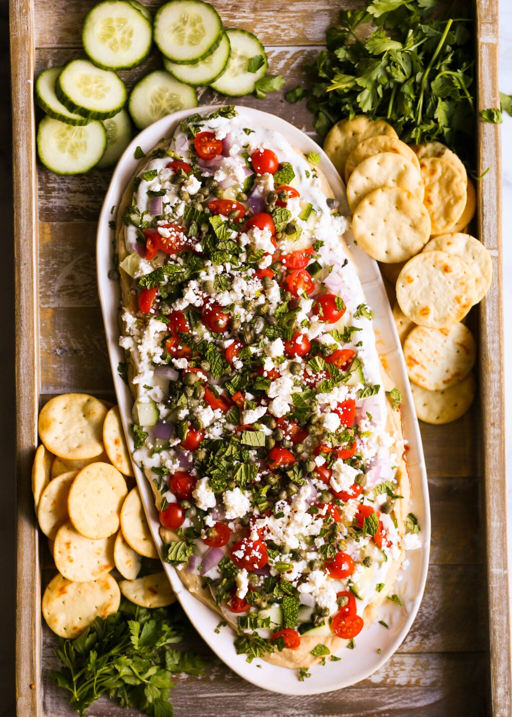 Layered Greek dip with hummus, tzatziki, cherry tomatoes, cucumbers, feta, and herbs, served with pita chips and cucumbers—easy Greek appetizer recipe.
