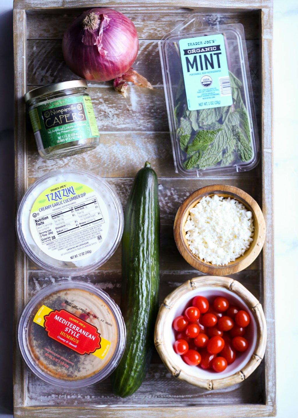 Fresh ingredients for a quick Greek layered dip recipe, featuring hummus, tzatziki, cucumber, feta, cherry tomatoes, red onion, capers, and mint.