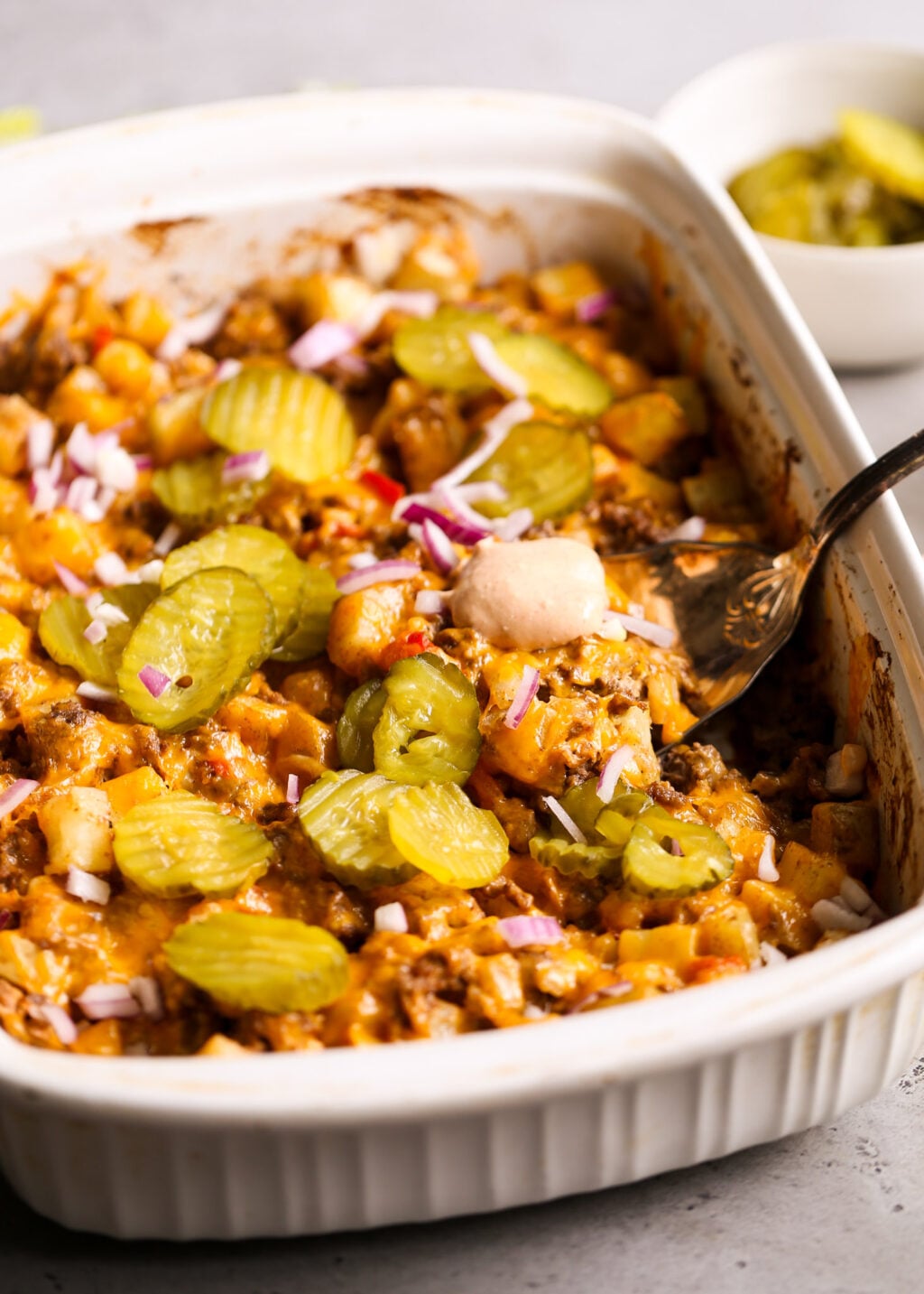 Cheeseburger casserole recipe with potatoes, ground beef, cheese, topped with pickle slices and red onions in a baking dish.
