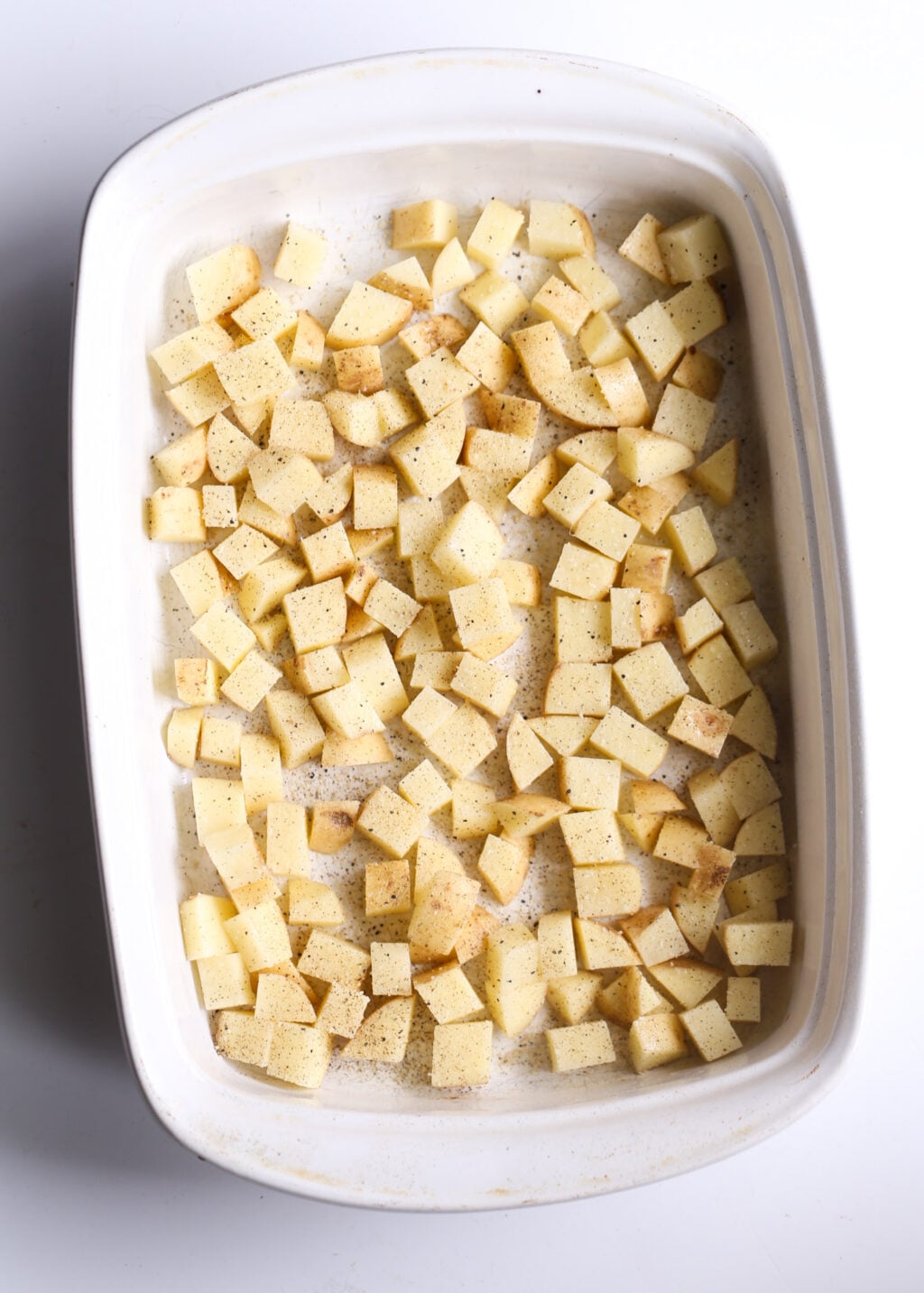 Creamy potatoes cubed and seasoned, spread in a white casserole dish, ready to bake for a cheeseburger casserole recipe.