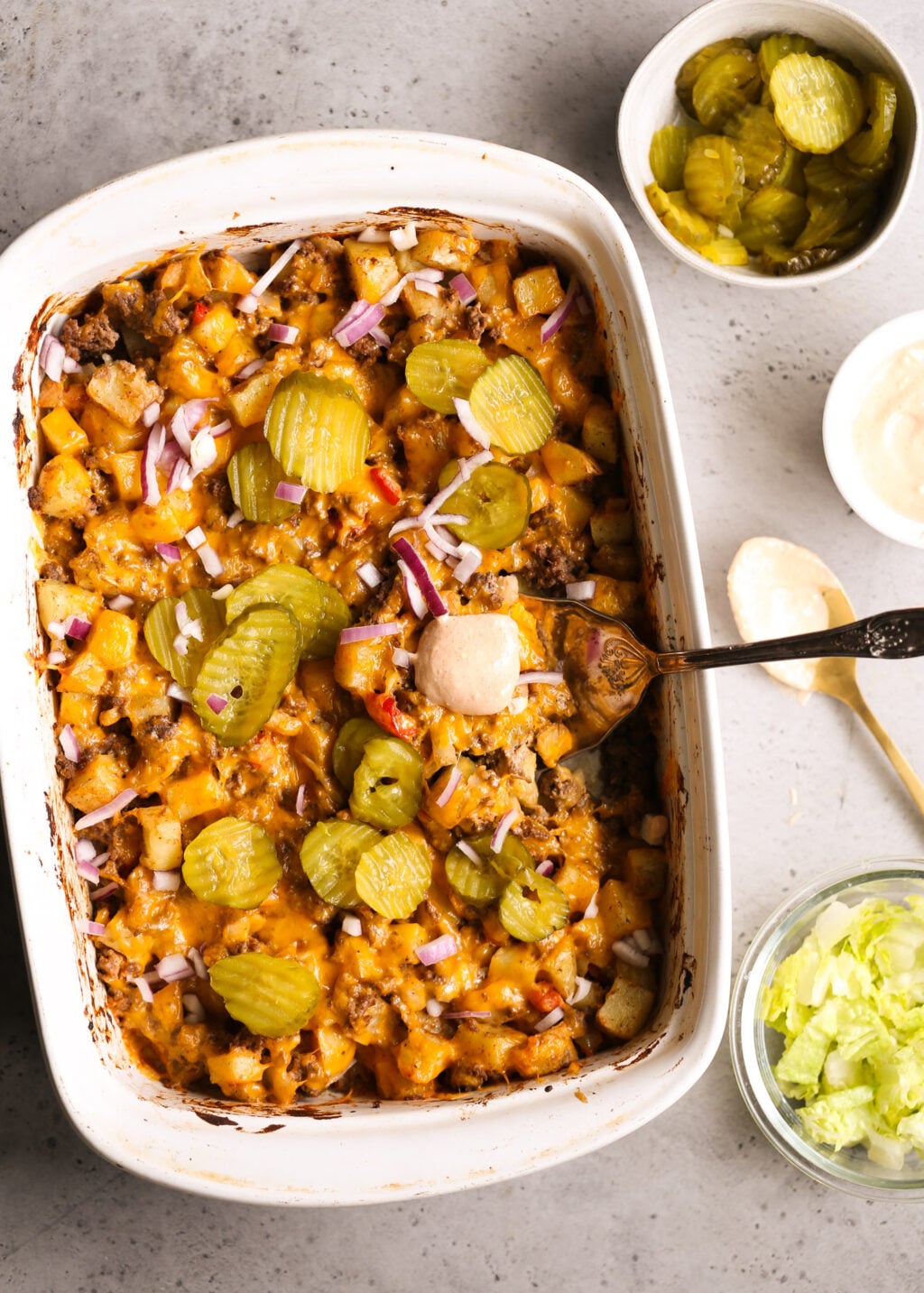 Cheeseburger casserole, baked, with potato cubes, ground beef, melted cheddar, topped with pickle slices and red onion.