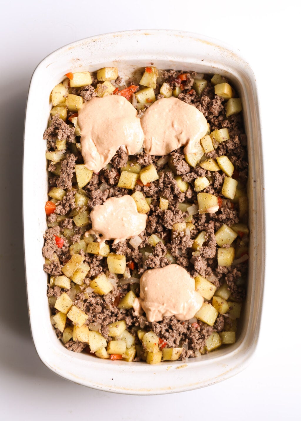 Creamy cheeseburger casserole with diced potatoes, ground beef, onions and peppers in a white baking dish, topped with dollops of sauce.