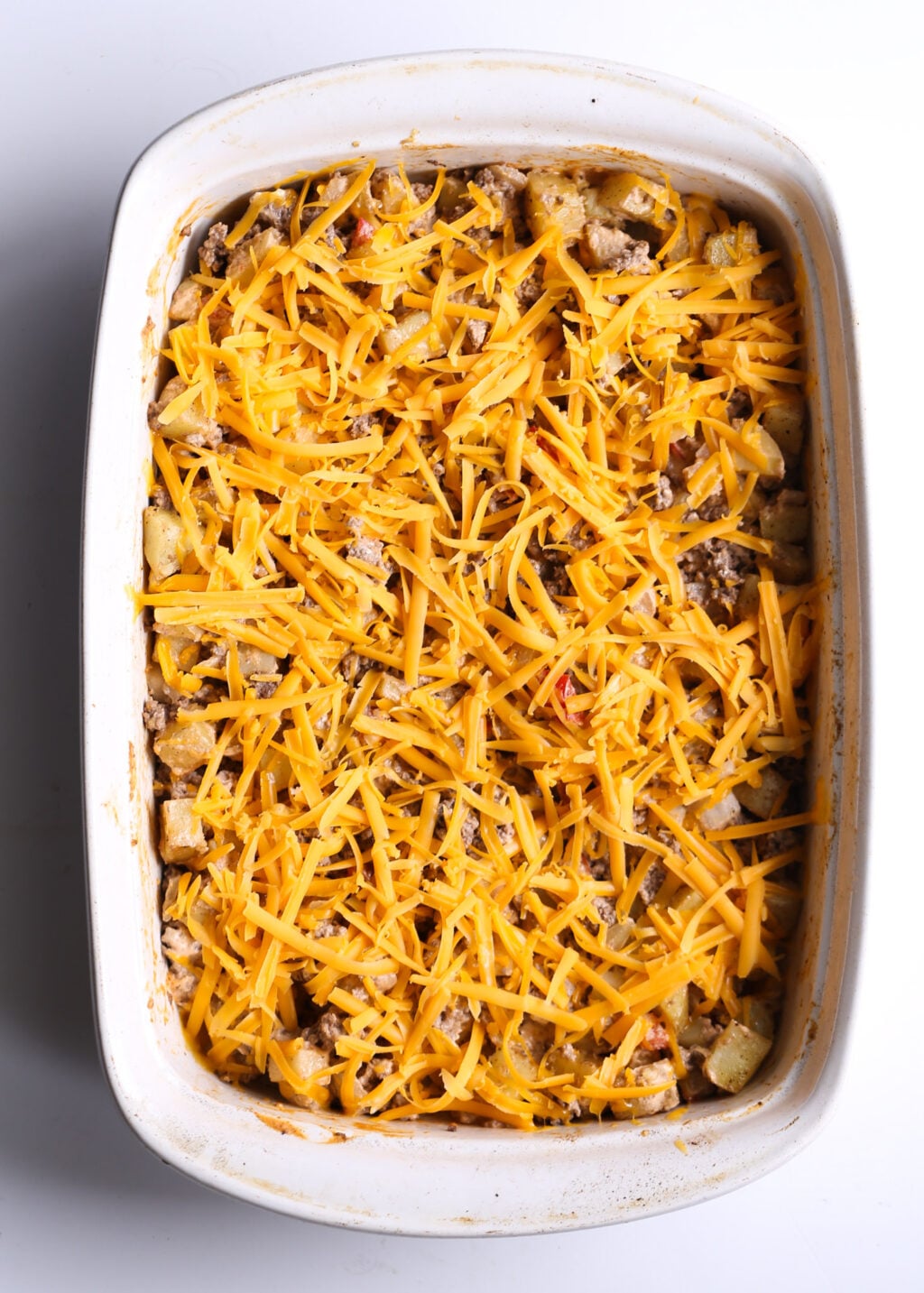 Cheeseburger casserole with potatoes, browned ground beef, and shredded cheddar on top, baked in a white casserole dish.
