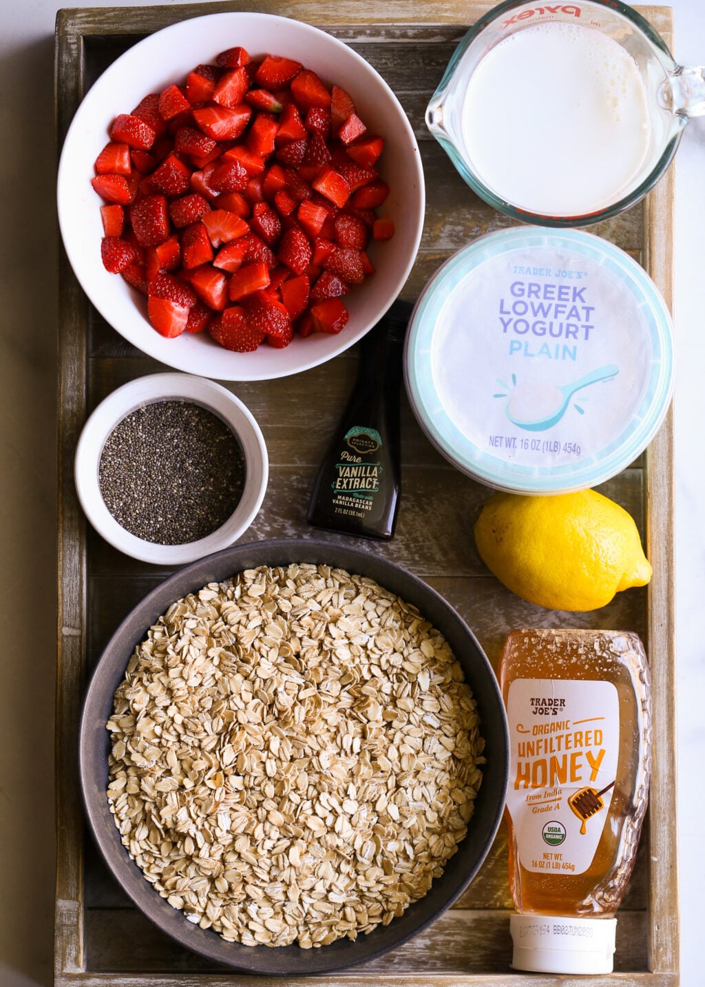 Fresh strawberries, oats, yogurt, milk, lemon, honey, vanilla, and chia seeds arranged on a wooden tray for strawberry-lemon overnight oats recipe.