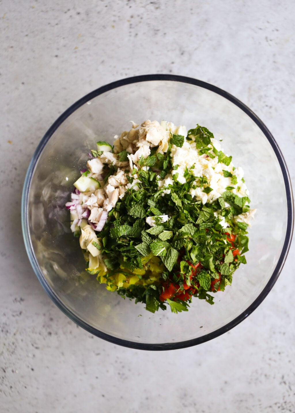Glass bowl of creamy Mediterranean chicken salad with combined prepped veggies, chicken, fresh herbs, and creamy base.